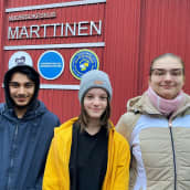 Kolme nuorta koululaista seisoo punaisen rakennuksen edessä hymyillen. Rakennuksen seinässä lukee Nuorisokeskus Marttinen. 