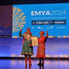 Two women in colourful traditional Sámi attire holding an award certificate and raising their fists on a stage.