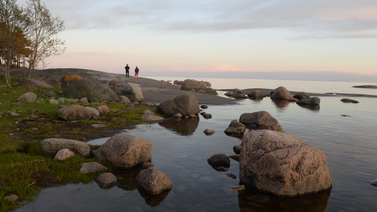 Kaksi ihmistä seisoo selin kivisellä luodolla. Etualalla on rantakiviä. On hieman hämärää ja taivaanranta on punertava.