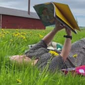 Nainen makaa selällään nurmikolla voikukkien ympäröimänä virkatulla värikkäällä peitolla. Hänellä on käsissään värikäs lastenkirja.