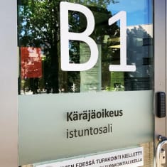 Talon ulko-ovi, jossa teksti B! ja sen alapuolella Käräjäoikeus istuntosali.
