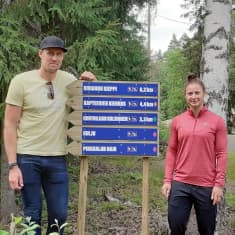 Marko Anttila vaaleankeltaisessa t-paidassa ja Jenni Hiirikoski vaaleanpunaisessa juoksutakissa seisovat uusien opaskylttien vieressä metsän siimeksessä.