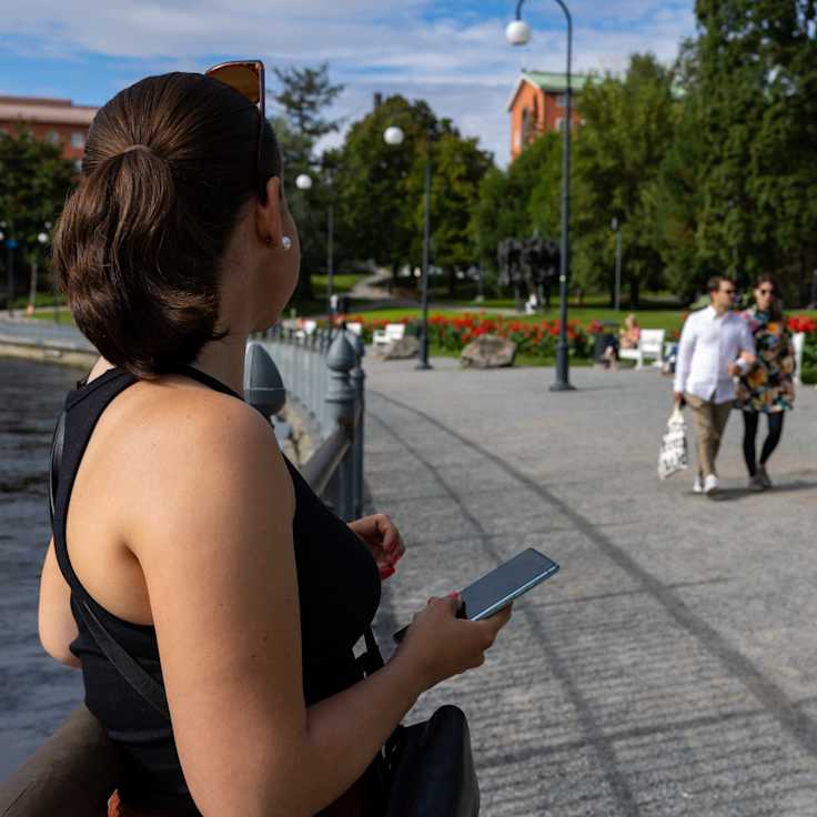 Anonyymi nainen puhelin kädessään katsoo kohti pariskuntaa Tampereen Koskipuistossa Tammerkosken rannalla.