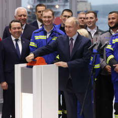 Vladimir Putin kääntääsuurta oranssia kytkintä. Taustalla sinisiin työvaatteisiin ja pukuihin pukeutuneita ihmisiä.