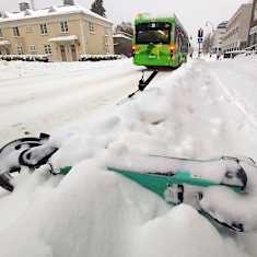 A green e-scooter on the ground in a snowbank, alongside a snowy street and sidewalk.