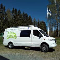 Kuvassa valkoinen pakettiauto, joka toimii Etelä-Savossa hyvinvointiautona. Kyljessä lukee vihreällä "Reissu-Ellu" eli auton nimi. Kuva on otettu keväällä.