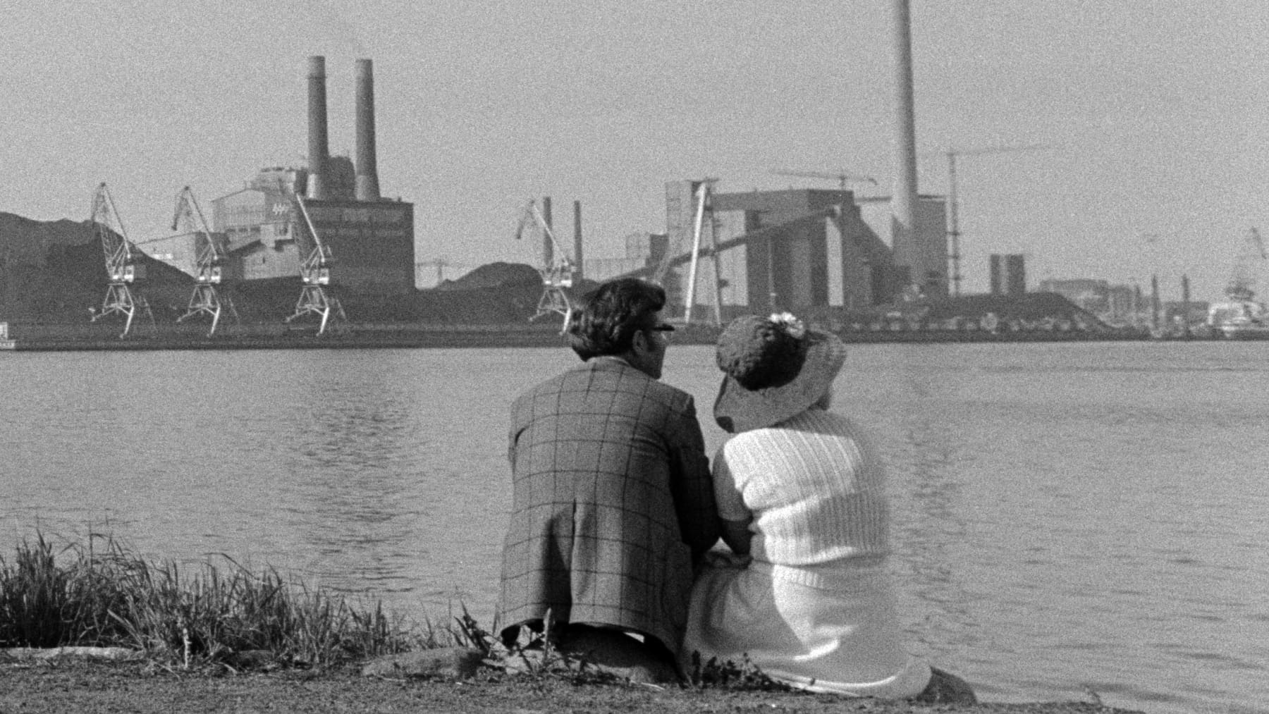 Mies ja nainen istumassa rantatöyräällä Tervasaaressa. Taustalla rakenteilla oleva Merihaka ja Hanasaaren voimalaitos.