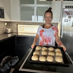 Entinen maajoukkuehiihtäjä Riitta-Liisa Roponen kotikeittiössään pitelemässä peltiä, jossa on vasta paistettuja sämpylöitä. Perheen koira Ibra istuu pöydän vieressä.