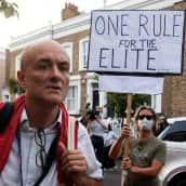 Punaisiin liiveihin ja valkoiseen paitaan pukeutunut Cummings kulkee  kadulla laukkua kantaen. Taustalla näkyy mielenosoittaja kyltin kanssa. Mielenosoittajalla on kasvomaski. Kyltissä on teksti: One rule for the elite..."