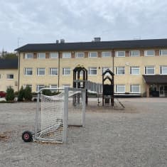 Vanha rappauspintainen vaaleankeltainen kolmikerroksinen koulurakennus, jonka pihassa on jalkapallomaali ja kiipeilyteline.