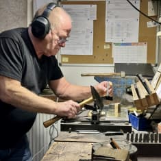 Vanha mies kuulosuojaimet korvillaan pitää vasaraa kädessään ja valmistautuu lyömään uuden rautaisen kirjaisimen metallilevyyn. Ympärillä työkaluja ja muistitauluja.