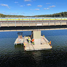 Workers in bright yellow and orange clothing seen beneath a concrete and steel bridge, with water surrounding them, and trees on the horizon.