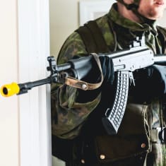 Maastopukuun pukeutunut mies pitelee rynnäkkkökivääriä, jonka piipussa sysäyksenvahvistaja.