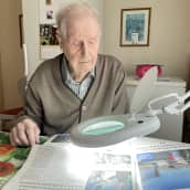 Vanha mies istumassa keittiön pöydän ääressä. Hän lukee sanomalehteä pöytään asennetulla suurennuslasilla.