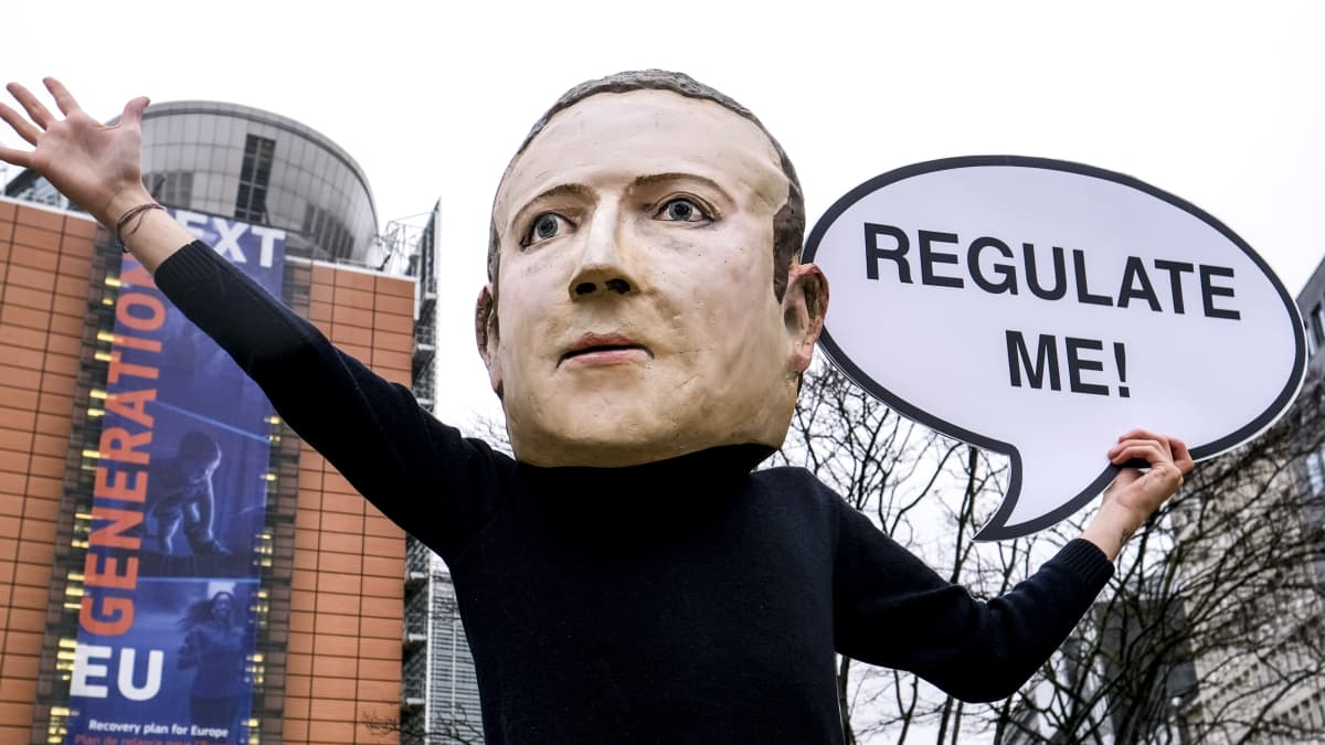 Mark Zuckerbergin näköinen maski kasvoillaan aktivisti osoittaa mieletään kädessää pahvinen puhekupla jossa lukee Regulate me.