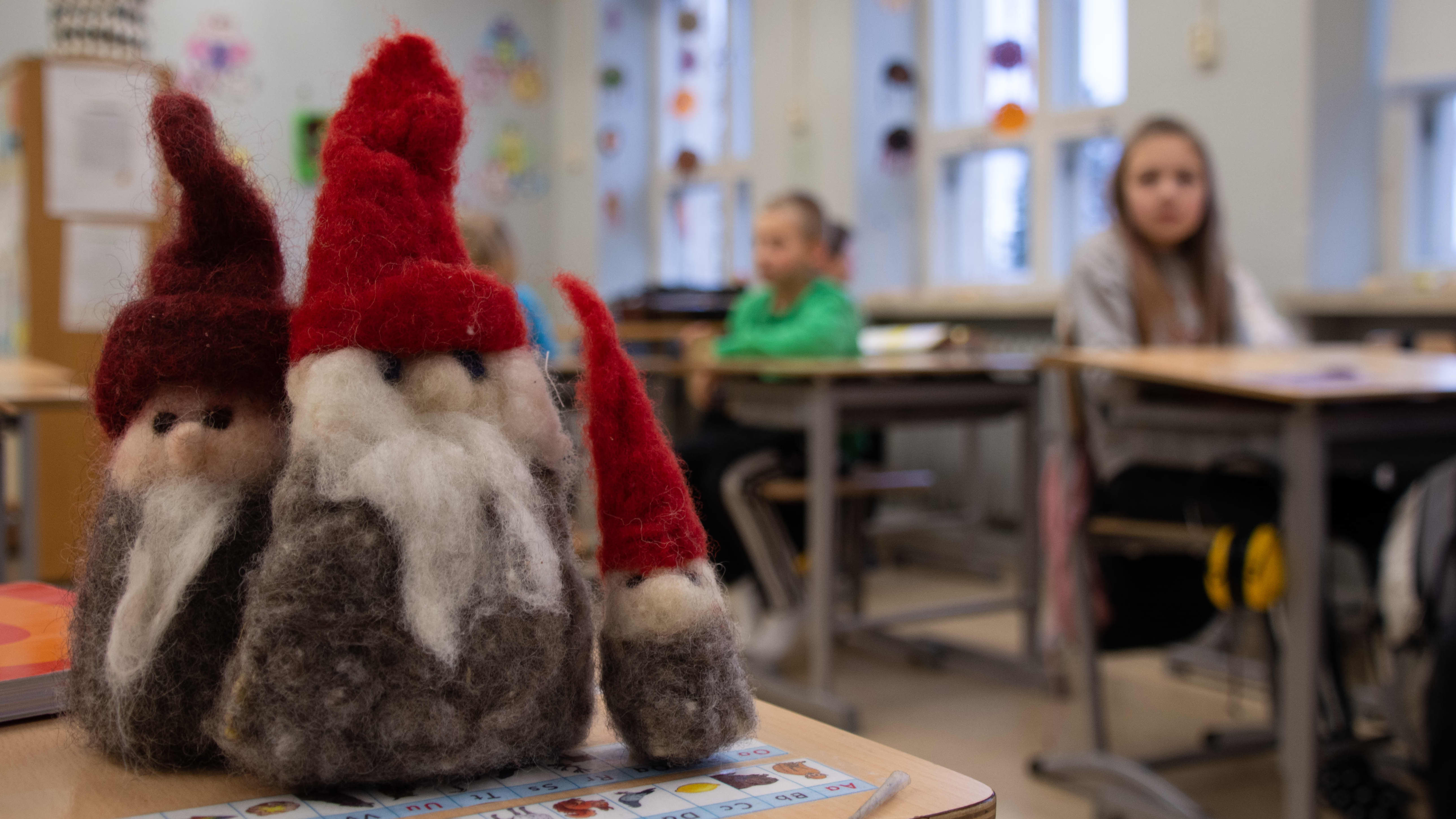 Tre tovade jultomtar står på en pulpet i ett klassrum, elever syns i bakgrunden.