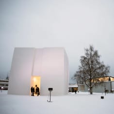 Lumesta rakennettu iso neliskanttinen kuutio, jonka keskellä on sisäänkäynti, kuution oviaukossa ihmisiä ja taustalla Rovaniemen pääkirjasto.
