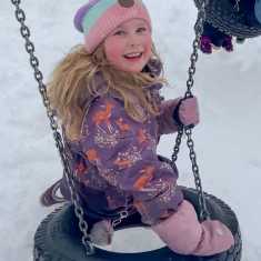 Pitkähiuksinen tyttö talvivaatteissa keinuu rengaskeinussa ja katsoo kameraan. Maassa on paljon lunta.