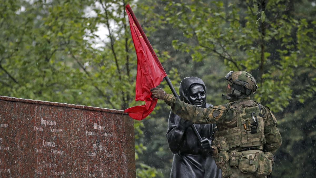 En soldat med ryggen till sätter en röd flagga på en mörkgrå staty av en kvinna.