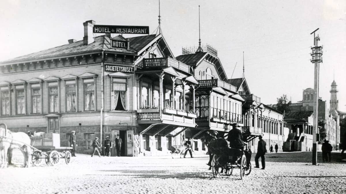 Mustavalkokuvassa puinen koristeellinen hotellirakennus, jonka edestä kulkee hevoskärryjä ja ihmisiä.