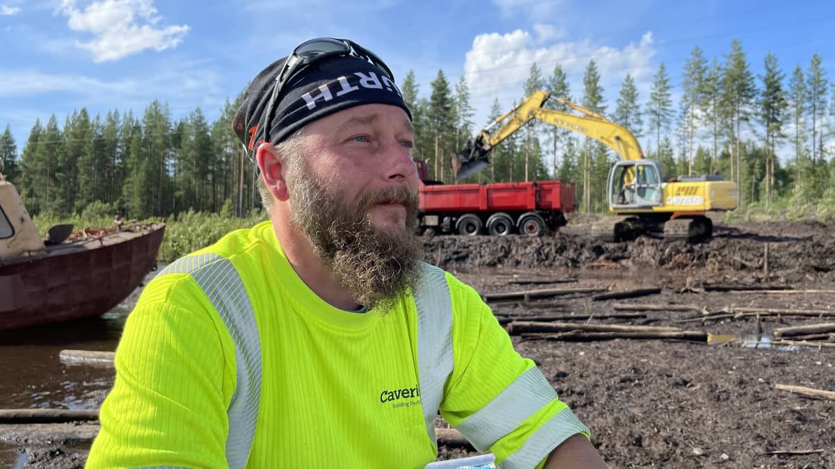 Mies istuu joen rannalla, taustalla näkyy turvetta, uppotukkeja ja kaivinkone.