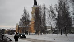 Talvinen katunäkymä, jossa nainen kävelee jalkakäytävällä ja tumma auto ajaa kadulla. Taustalla koivuja ja keltainen iso Oulun tuomiokirkko.