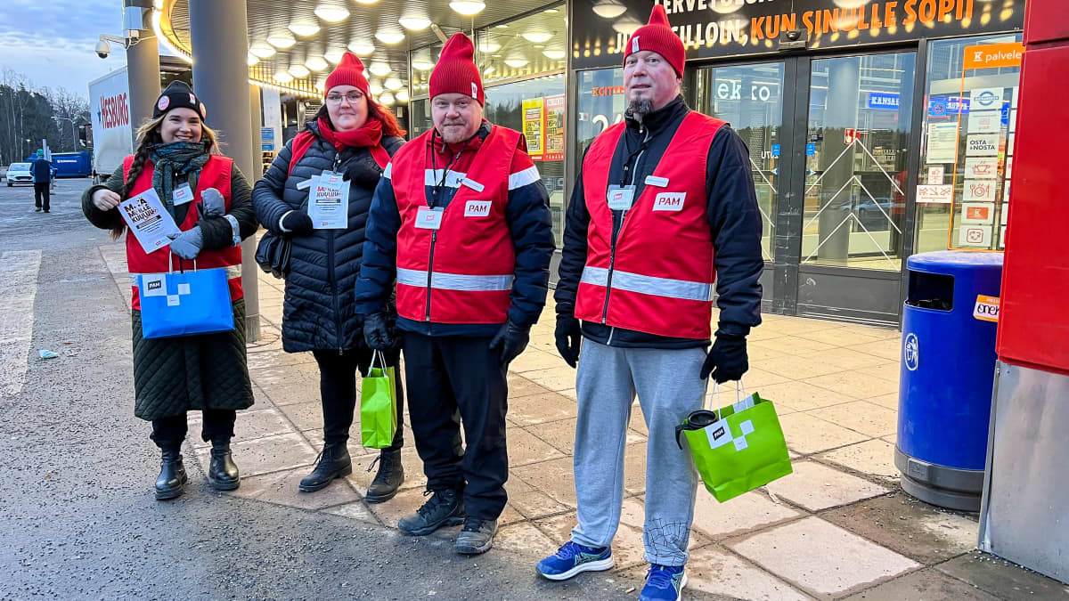 Neljä lakkovahtia, osa punaisissa liiveissä suuren marketin edustalla.