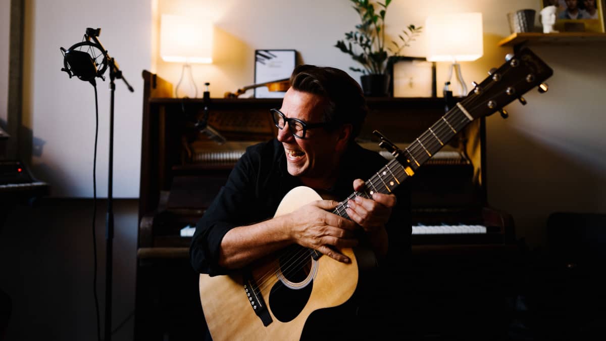 Mikko Kuustonen istuu äänitysstudiolla kitara sylissään ja nauraa.