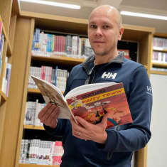 Professori Mika Rämet kirjaston sarjakuvaosastolla kädessään Lucky Luke -sarjakuva.