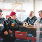 Neljä eläkeläismiestä istuu huoltoaseman pöydässä kahvilla ja keskustelee hymyillen.
