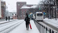 Person walking along a snow-covered tram platform on a city street, with falling snow, buses and a few other people in the background.