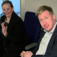 Mies virnistää kameralle tulkkauskopista, ja taka-alalla nainen ojentaa miehelle kuulokkeet.