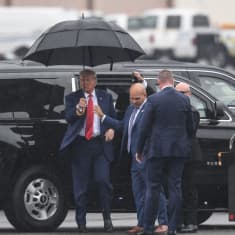 Yhdysvaltain entinen presidentti Donald Trump nousee mustasta tila-autosta lentokentällä ja pitelee mustaa sateenvarjoa.