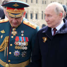 Sergei Šoigu ja Vladimir Putin kävelevät Punaisella torilla. Šoigulla on tummanvihreä juhlaunivormu arvomerkkeineen ja koppalakki, Putinilla musta päällystakki, punainen kravatti ja rintapielessään oranssi-musta yrjönnauha. Taustalla näkyy häimeänä tavaratalo GUM.