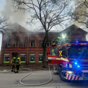 Punatiilinen rakennus palaa. Palomiehiä sammuttamassa. Paloauton valot vilkkuvat.