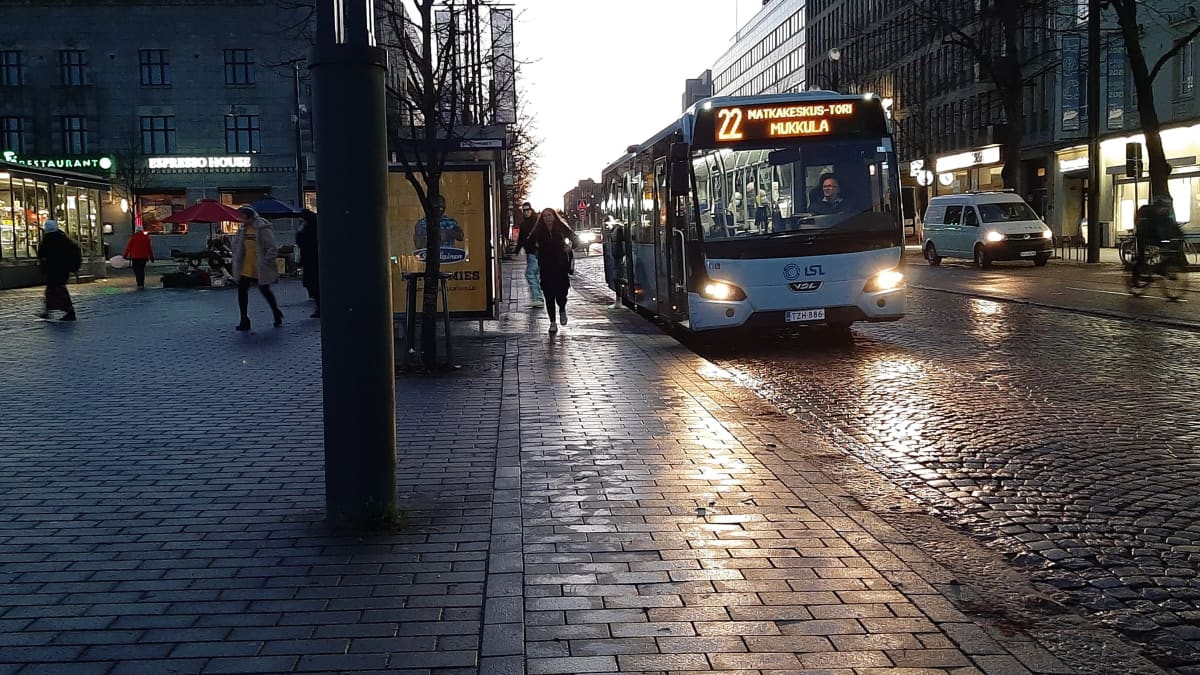 Paikallisliikenteen sininen bussi aamuhämärissä, bussista ulos tulevia matkustajia, katu kiiltää kosteudesta bussin valoissa.