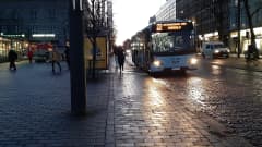 Paikallisliikenteen sininen bussi aamuhämärissä, bussista ulos tulevia matkustajia, katu kiiltää kosteudesta bussin valoissa.