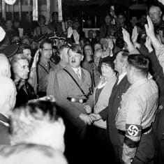 Adolf Hitler kanttajiensa keskellä kuvattuna. Takana on hymyileviä ja hailaavia natsipukuisia miehiä. 