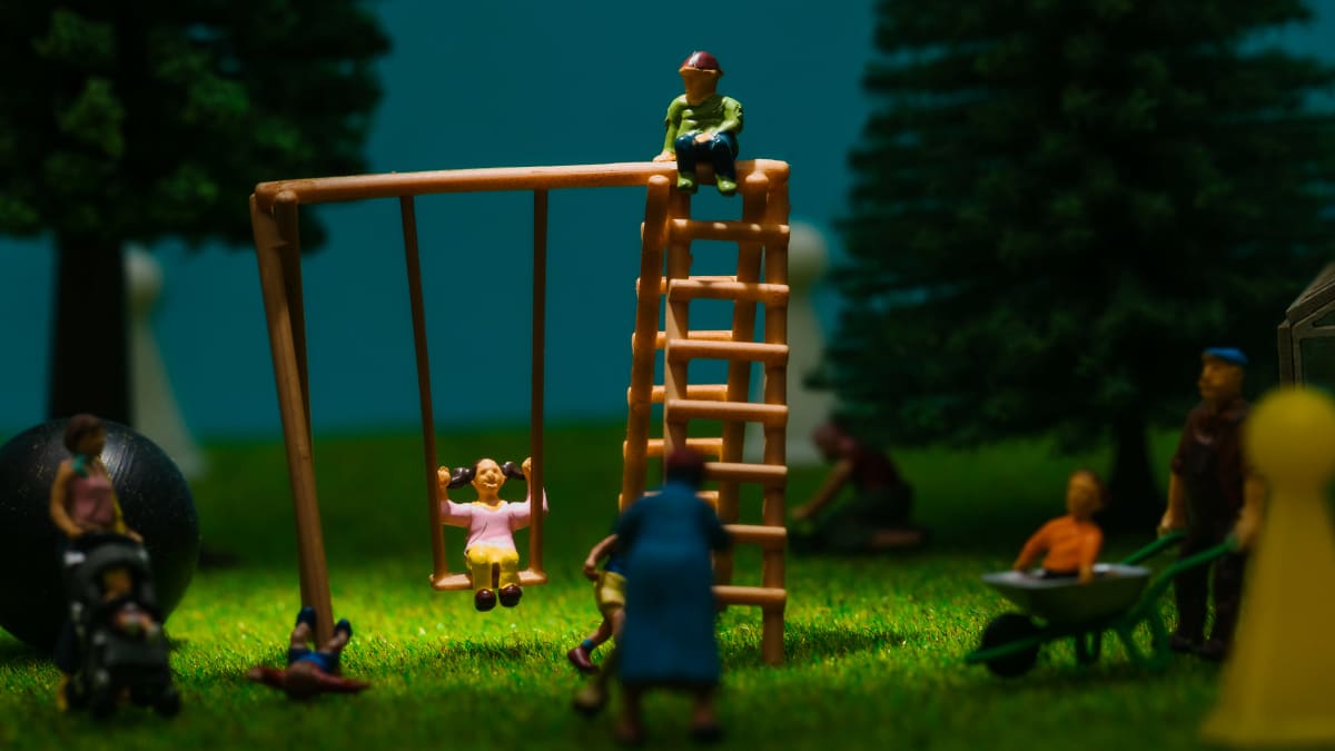 Miniatyyrimaisema leikkipuistosta. Lapsia leikkii keinussa, yksi lapsi on kaatunut, äiti lastenvaunujen kanssa katsoo tilannetta.