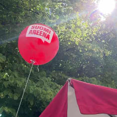 Punainen ilmapallo jossa lukee SuomiAreena leijuu vihreälehtisen puun edessä. Aurinko paistaa oksien läpi.