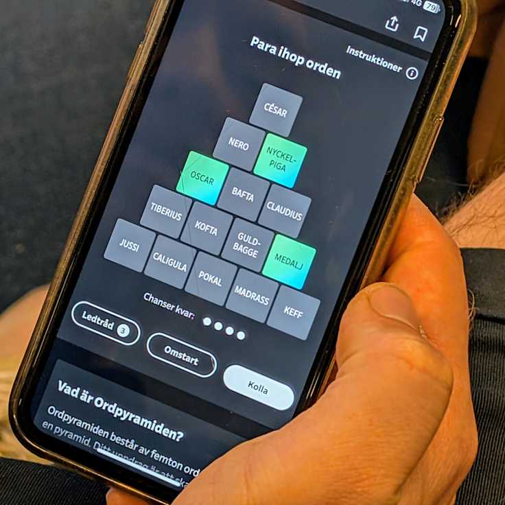 En person håller en smartphone som visar ett ordpussel på skärmen. Uppgiften är att para ihop olika ord.