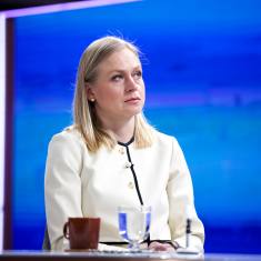 Elina Valtonen, with shoulder-length, straight blonde hair, wearing a white jacket, sitting at a table in front of a black microphone, glass of water and brown mug, with a blue background.