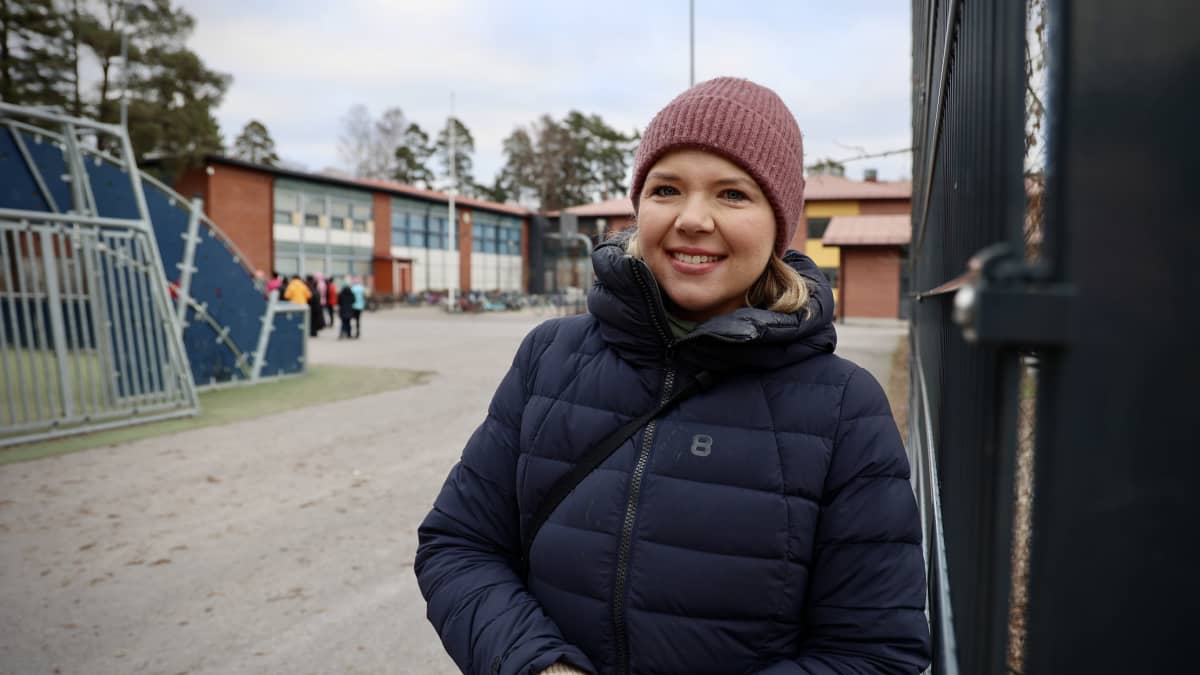 Nainen hymyilee kameralle, taustalla näkyy koulurakennus ja oppilaita.