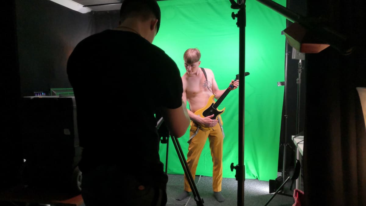Mies poseeraa kitaran kanssa kuvausseinän edessä. Toinen ottaa valokuvaa. 