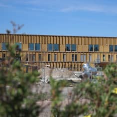 Rakennustyömaa-aitojen takana näkyy rakenteilla oleva ruskea kaksikerroksinen laitosrakennus.