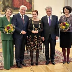 Three formally dressed women and two older men in grey suits standing in a row.