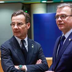 Two men with dark blue suits and glasses stand with serious expressions in a meeting room.