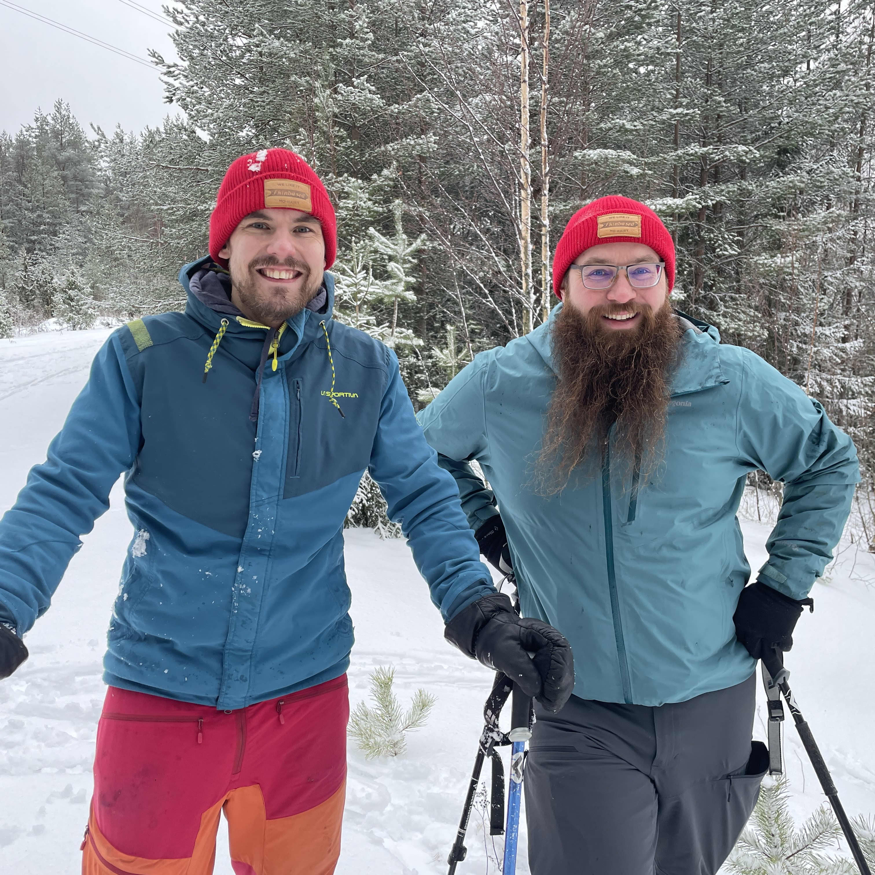 Kaksi iloisesti hymyilevää miestä suksilla lumisessa metsässä. Punaiset pipot päässä, molemmilla sinisävyiset takit.
