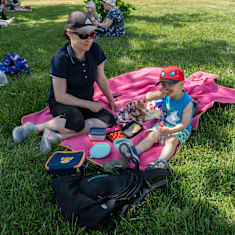 En kvinna och ett barn sitter på en rosa piknikfilt i gräset.