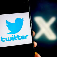 En hand håller upp en telefon med Twitters logo, i bakgrunden syns ett X.